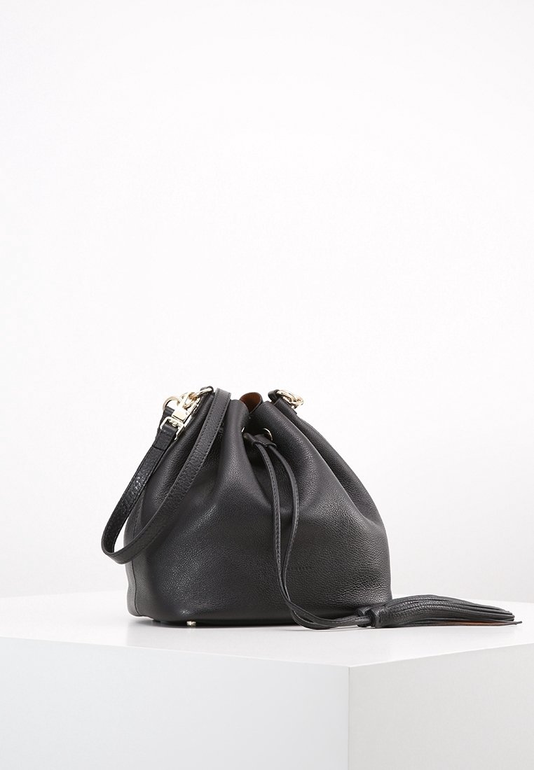 Sac seau en cuir noir avec fermeture à cordon et bandoulière, posé sur une surface blanche sur fond blanc uni.