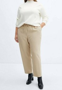 Beige weit geschnittene Hose mit elastischem Bund und Kordelzug, kombiniert mit einem crèmefarbenen Langarmoberteil. Schwarze Stiefeletten mit Absatz vervollständigen das Outfit.