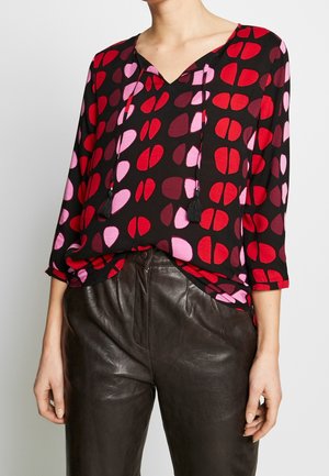 Femme portant un chemisier noir avec des formes abstraites rouges et roses et un pantalon en cuir noir, debout sur un fond neutre.