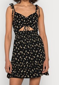Svart blommig klänning med justerbara knytband, rynkad byst, utskärning och utsvängd volangkant. Har små gula och blå blommor.