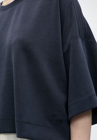 Chemise bleu marine foncé à manches courtes avec détails côtelés aux épaules et logo brodé discret près de l'ourlet, portée sur un fond clair.
