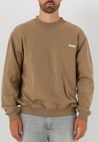 Brun crewneck sweatshirt i bomullsblandning med ribbade muddar och nederkant. Litet vitt logotyp "WOODBIRD" på vänster bröst.