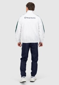 Weißes Jackett mit grünen Akzenten, elastischen Bündchen und Logo auf dem Rücken, kombiniert mit navyfarbenen Hosen. Glattes, leichtes Material und sportliches Design.