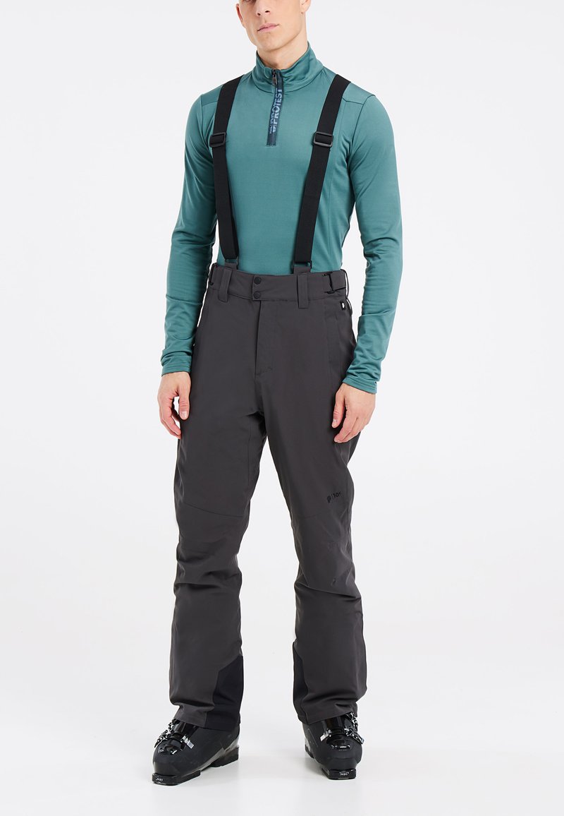 Zwarte skibroek met verstelbare schouderbanden, met een recht model. Gecombineerd met een groene lange mouwen top en zwarte skischoenen.