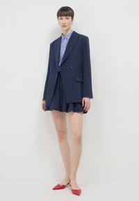 Blazer navy su una camicia a quadri blu, abbinato a una gonna svasata navy e scarpe rosse a punta, con un design su misura e linee pulite.