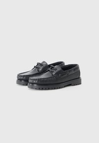 Sorte lær båtsko med moccasin-stil foran, gummisåle med mønster, og snøring nær kragen. Jevn tekstur gjennom hele.