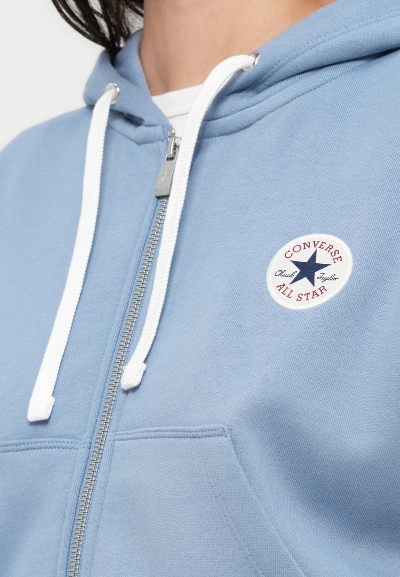 Sweat à capuche zippé bleu clair avec cordons blancs ; présente un patch rond avec une étoile et le texte "Converse All Star". Texture douce.