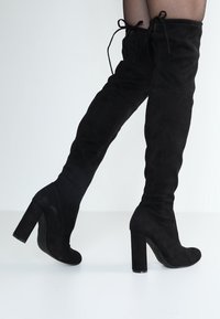 Schwarze Wildlederstiefel über das Knie mit hohem Blockabsatz und Schnürdetails an der Oberseite. Glatte Textur und figurschmeichelnde Silhouette.