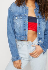 Mujer con chaqueta de mezclilla azul claro sobre una blusa bandeau roja y azul marino y jeans azul claro, con la mano en la cadera.