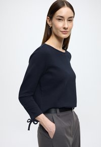 Granatowa teksturowana bluzka z długim rękawem i okrągłym dekoltem oraz sznurkami przy mankietach, zestawiona z szarymi, wysokim stanem spodniami i czarnym paskiem.