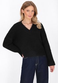 Schwarzer, flauschiger Strickpullover mit V-Ausschnitt und weiten Ärmeln. Der Saum ist kurz geschnitten. Kombiniert mit dunkelblauen Jeans aus Denim mit hellen Nähten.