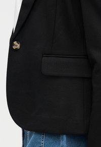 Gros plan sur un blazer noir avec un bouton en écaille de tortue et une poche à rabat, porté avec un jean en denim bleu.