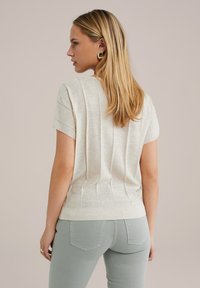 Gai bēšs adīts tops ar īsām piedurknēm, ar vertikālu ribu apdari un pieguļošu apakšu. Apvienots ar gaiši zaļām pieguļošām džinsām.