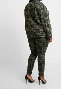 Camo-patterned jacket and pants in varying shades of green, paired with black high heels. Fabric appears smooth and fitted.