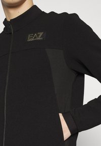 Giacca nera in maglia con tessuto testurizzato, chiusura con zip e patch del logo. Presenta pannelli neri lisci e polsini a coste per un dettaglio aggiuntivo.