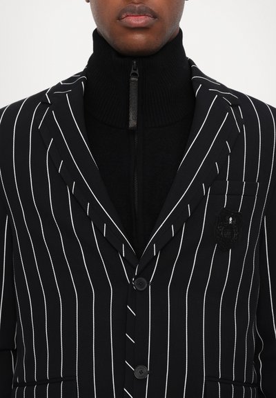 Black pinstriped blazer with a black zipper neck underneath. Features two buttons, a chest pocket, and a textured black emblem.