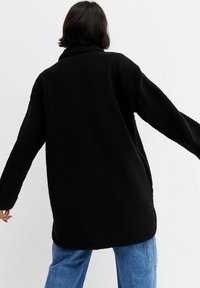 Chaqueta de forro polar negra con un corte relajado, mangas largas y un cuello clásico, combinada con unos vaqueros de pierna ancha en azul claro.