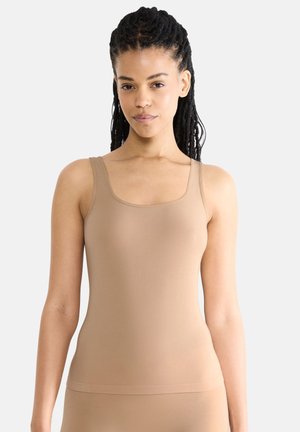 Young woman with braided hair wearing a fitted beige sleeveless top, standing against a plain light background.