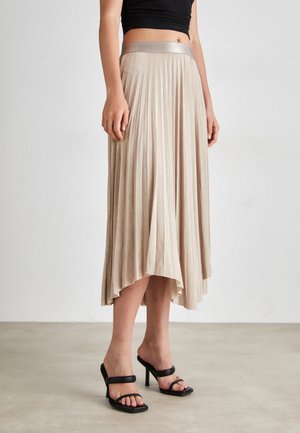 Vrouw draagt een beige geplooide midirok met een asymmetrische zoom en zwarte sandalen met hoge hakken, staand op een effen vloer.