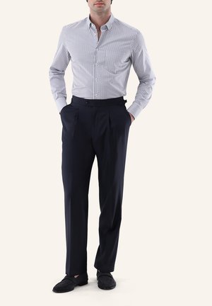 Mann trägt schwarze plissierte Hose, schwarze Loafers und ein weißes Hemd mit kleinem schwarzem Karo-Muster, Hände in den Taschen.