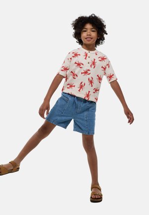 Chemise blanche à manches courtes avec un imprimé de homard rouge, associée à un short en denim bleu. L'enfant porte des sandales beiges et adopte une pose ludique.