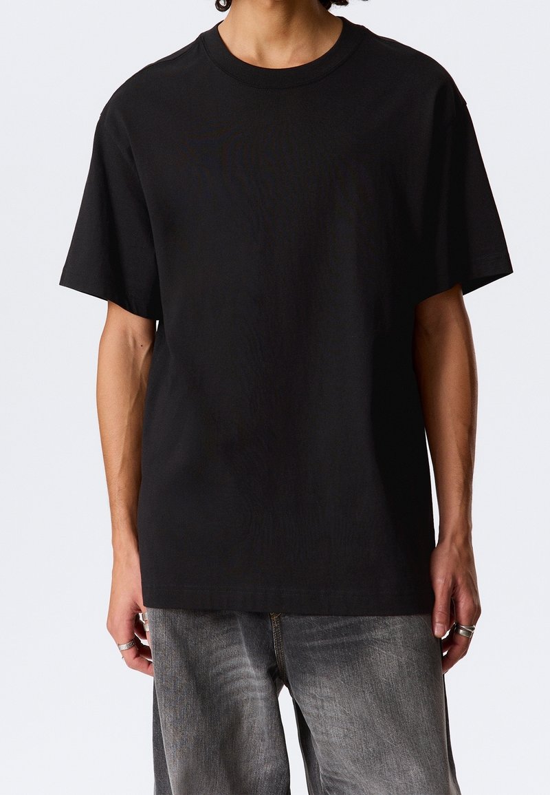 Personne portant un t-shirt noir uni oversize à manches courtes et un jean gris délavé, debout les bras détendus sur les côtés.