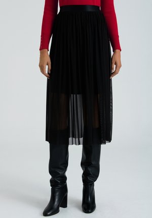 Woman wearing a black pleated midi skirt with sheer overlay, red long-sleeve top, and black knee-high heeled boots, standing against plain background.