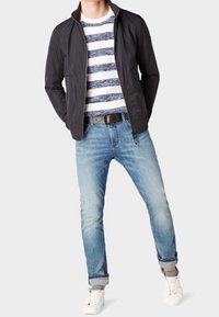 Giacca leggera nera, camicia a righe blu e bianca, jeans blu chiaro con orli risvoltati e sneakers bianche. Un outfit semplice e informale.