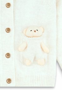 Cárdigan de punto color crema con botones de madera y un bolsillo que presenta un pequeño detalle de peluche de oso en beige claro, con una textura suave.