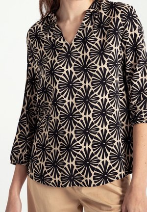 Kvinde iført beige bluse med sort abstrakt blomsterprint og trekvartlange ærmer, kombineret med beige bukser.