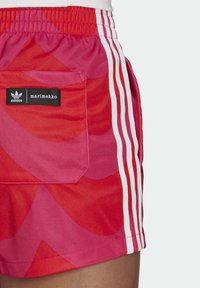 Red and pink shorts featuring a bold pattern, elastic waistband, side pockets, and white adidas stripes along the sides. Black logo tag present.