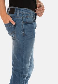 Jean en denim bleu coupe droite, avec un design à cinq poches, des coutures visibles et une étiquette en cuir sur la taille.