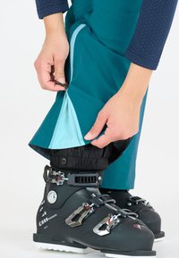 Tealfärgade vattentäta skidbyxor med en blå innervadding, som har en dragkedja vid ankeln och justerbar elastisk mudd över svarta skidstövlar.