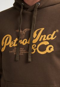 Bruine hoodie met een grote, gestructureerde gele opdruk van "Petrol Ind & Co" en een adelaar embleem. Inclusief verstelbare trekkoorden.