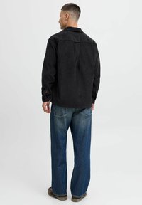 Chaqueta de pana negra con un ajuste relajado, que presenta un yugo en la espalda, mangas largas y puños abotonados, combinada con unos jeans de mezclilla azul de corte holgado.