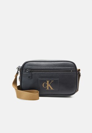 Schwarze Leder-Crossbody-Tasche mit vorderer Reißverschlusstasche, goldenem „cK“-Logo und verstellbarem braunem Stoffriemen vor weißem Hintergrund.