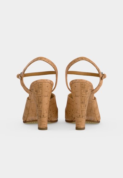 Ζευγάρι πέδιλα με υφή φελλού, ψηλά τακούνια, λουράκια στον αστράγαλο και χοντρά τακούνια, εμφανίζονται από πίσω σε λευκό φόντο.