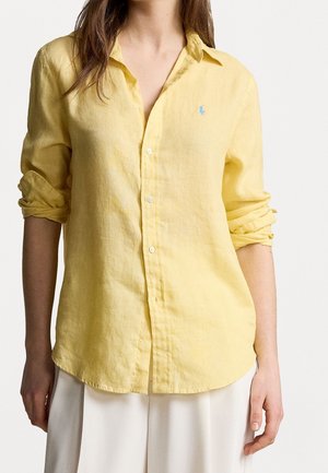 Femme portant une chemise en lin jaune clair boutonnée avec manches retroussées et pantalon blanc, visible des épaules aux hanches.