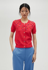 Cardigan court à manches courtes rouge avec des perles décoratives multicolores, col rond et fermeture à boutons ; porté avec un pantalon bleu.