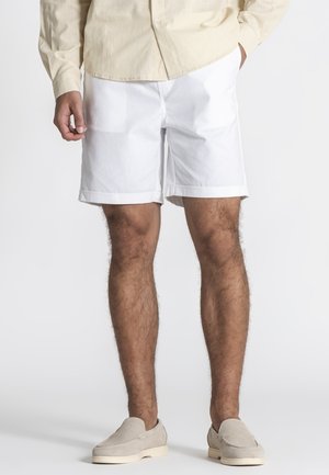 Mann trägt weiße, maßgeschneiderte Shorts, beiges Langarmhemd und beige Slip-On-Schuhe, steht mit einer Hand in der Tasche vor weißem Hintergrund.