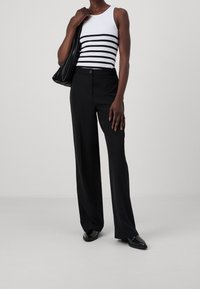 Henkilö, joka pukeutuu mustiin leveisiin housuihin, mustiin kenkiin ja valkoiseen hihattomaan paitaan, jossa on mustia vaakaraitoja, pidetään mustaa laskostettua olkalaukkua.