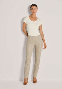 Vrouw in een crème kleurige top met korte mouwen, beige patroon broek en bruine hakken, staande tegen een lichtgrijze muur.