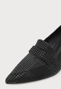 Mocassin noir à bout pointu avec motif pied-de-poule texturé et brillant et une large bride sur le dessus.