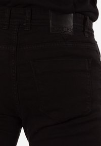 Vaqueros de mezclilla negros con etiqueta de cuero en la cintura. Cuenta con un bolsillo trasero con un detalle sutil de costura. El material parece liso y resistente.