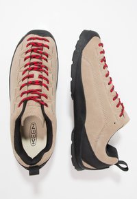 Beige suede hiking shoes with red laces, black rubber soles, and a black mesh accent. Features a pull tab at the heel for easy wear.