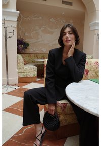 Musta räätälöity puku, jossa on istuva takki ja leveälahkeiset housut, yhdistettynä kudottuun mustaan käsilaukkuun. Taustalla marmoripöytä ja kukkakuosiset verhoilut.