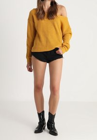 Överdimensionerad senapsgul stickad tröja med avskuren axeldesign, kombinerad med svarta denimshorts och svarta ankelstövlar.