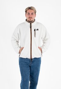 Biała polarowa kurtka z wysokim kołnierzem, pomarańczowym i czarnym zamkiem oraz kieszenią na lewej piersi. Połączona z niebieskimi dżinsami.