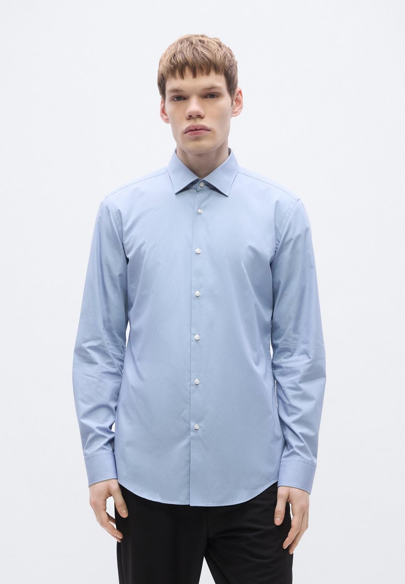 Helehallid särgipluus, mis on valmistatud siledast kangast, klassikalise krae ja nööpidega esiküljel. Lõige on keha järgi kohandatud, ümarate äärtega.