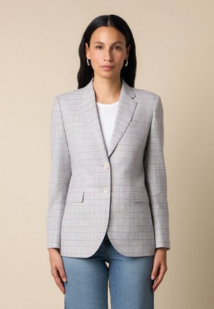 Vrouw draagt een lichtgrijze geruite blazer over een wit topje en blauwe spijkerbroek, staand tegen een effen beige achtergrond.